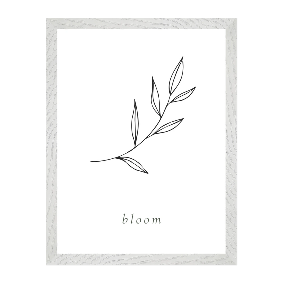 A framed minimalist art print featuring a line drawing of a leafy branch on a white background, with the word 'bloom' printed below in a small serif font. The frame is a light, textured white wood.